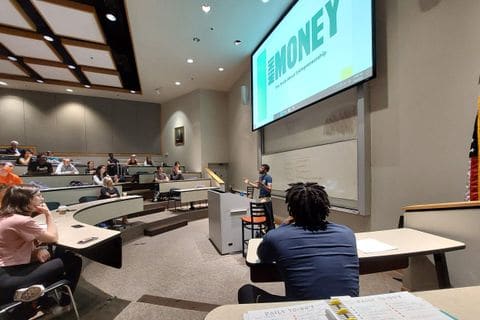 A lecture hall seen from the back row, a Mini Money slide on the screen at the front.