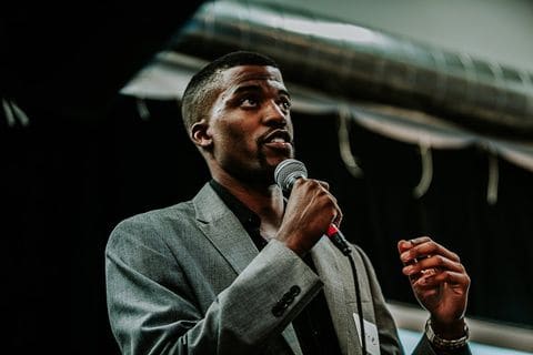 A speaker in a grey jacket addressing the room with a microphone at a Mini Money event.